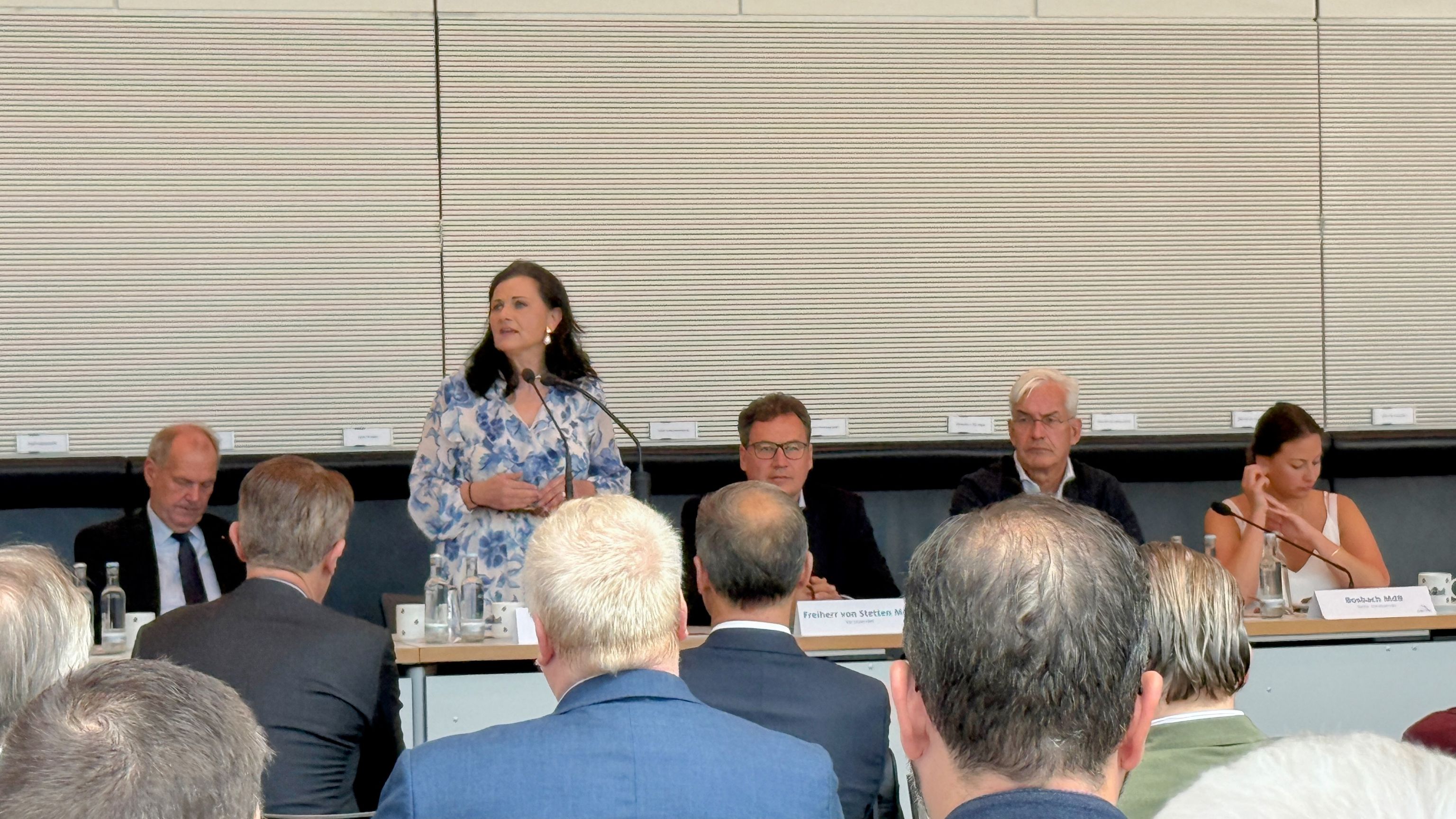 Frau hält Vortrag vor Publikum an einem runden Tisch mit Diskussionsteilnehmern.