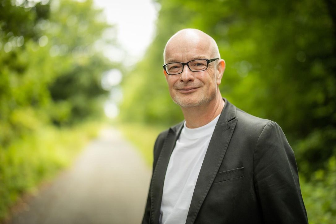 Bald glatzköpfiger Mann mit Brille lächelt in grünem Waldumfeld.