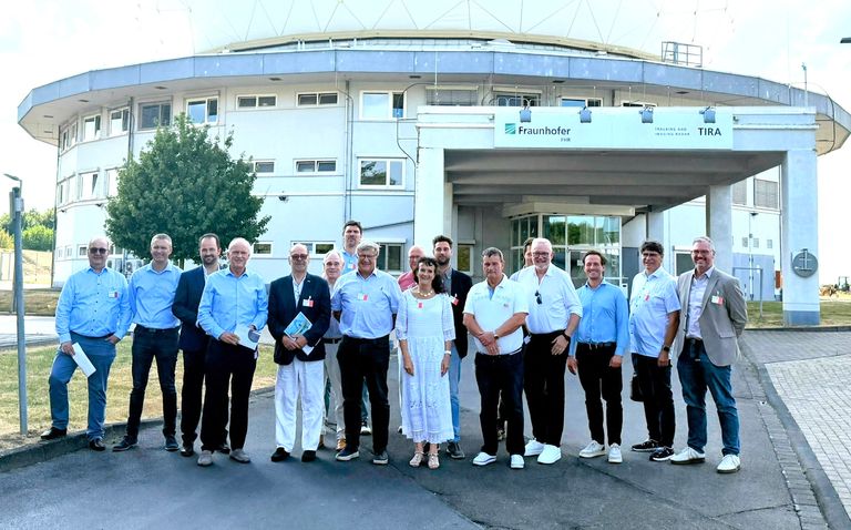 Gruppe von 14 Personen vor modernem Gebäude, alle lächeln in die Kamera.