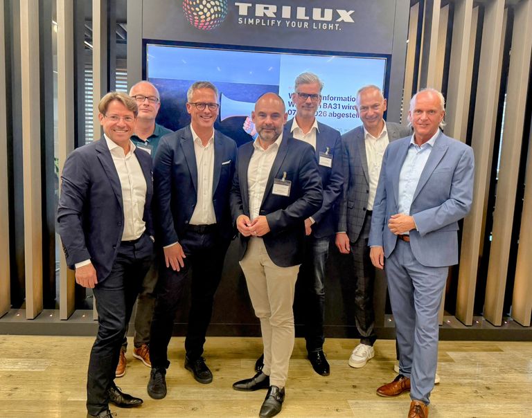 Sechs Herren in Businesskleidung vor Trilux-Wand mit Logo „Simplify Your Light“.
